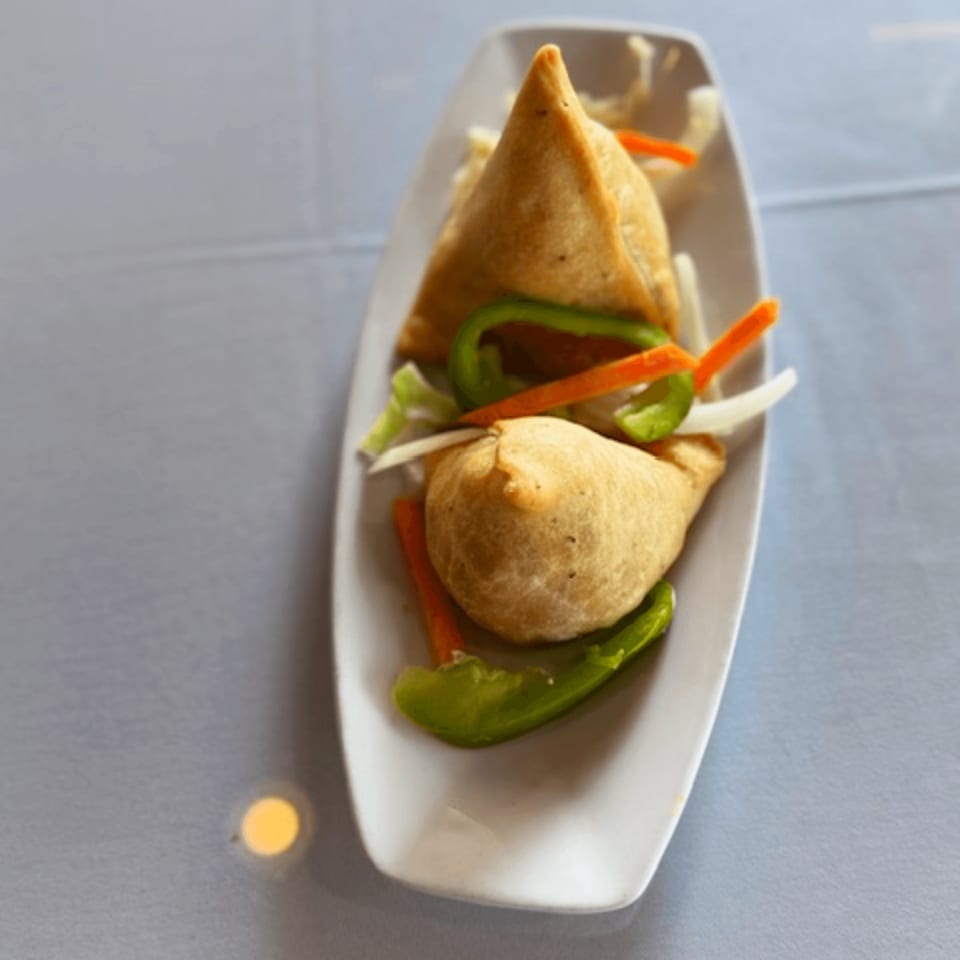 Two golden-brown Indian samosas garnished with sliced green peppers, carrots, and onions on a white plate.