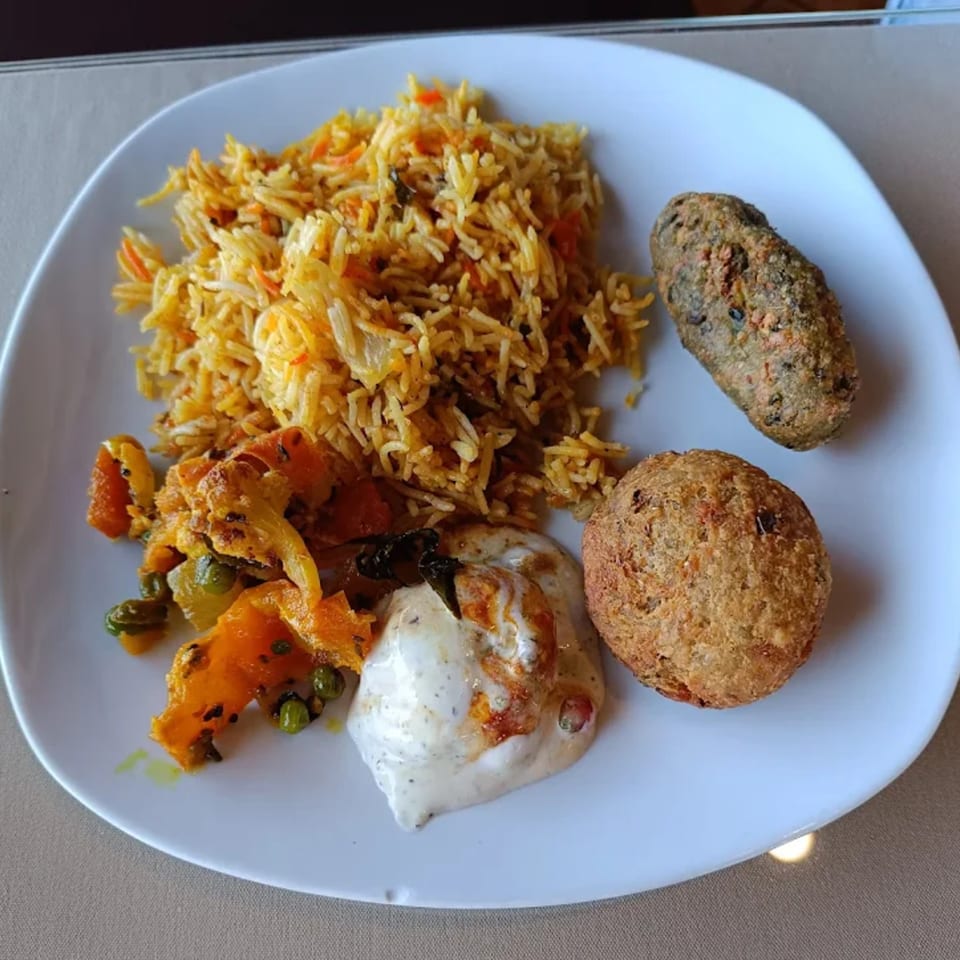 A white plate of Royal India Restaurant's Indian food, including biryani, vegetables, two fried cutlets, and raita.